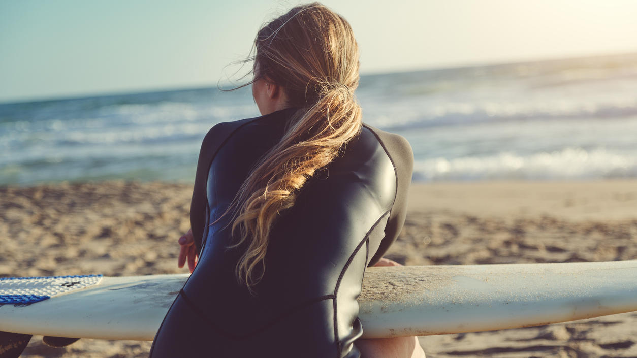 Fashion Lexikon: Neopren Surferin sitzt im Anzug aus Neopren am Strand