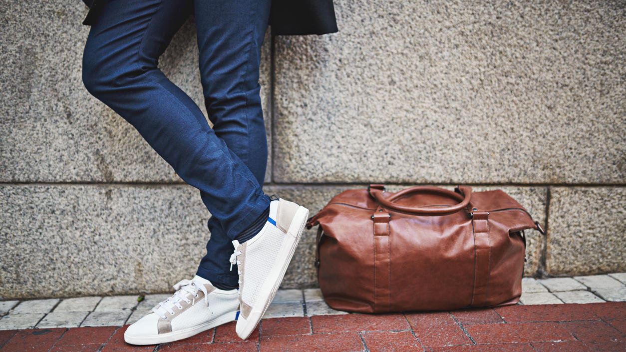 Fashion Lexikon: Weekender Mann in Jeans und Sneakern lehnt mit seinem braunen Weekender gegen eine Steinwand