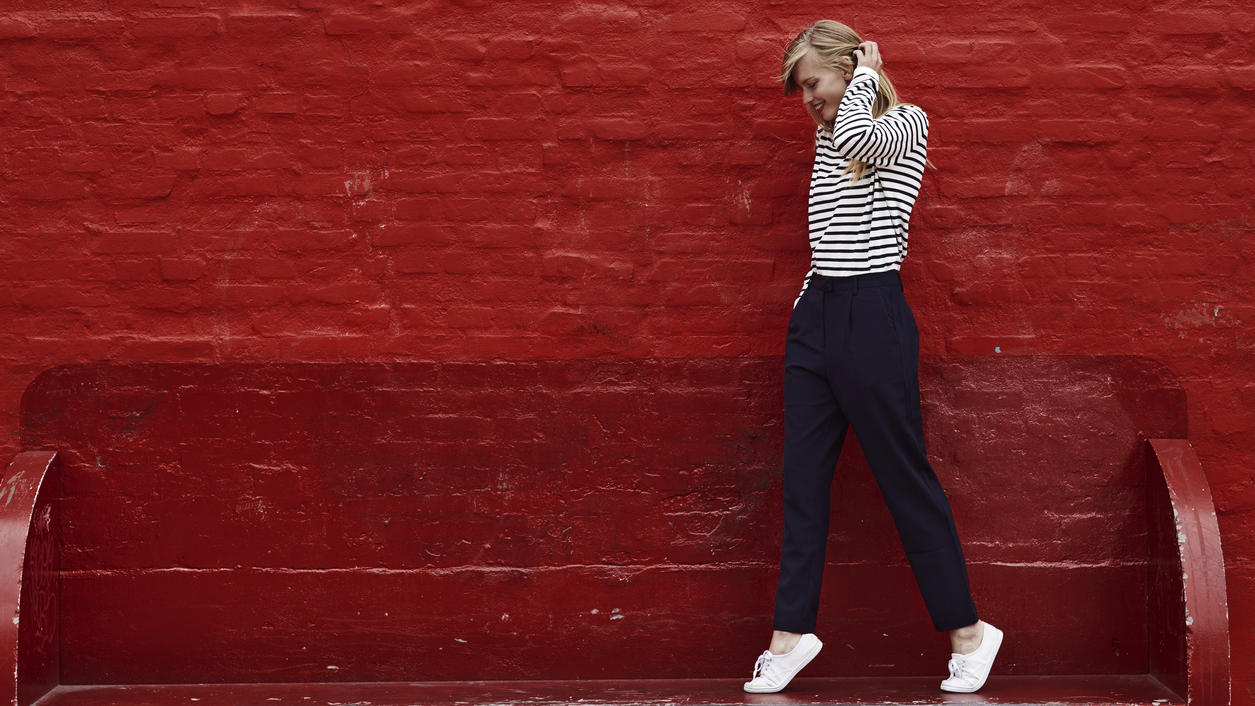 Fashion Lexikon: Loose Fit Junge Frau steht vor einer roten Wand und trägt eine Loose Fit-Hose mit einem weiß-schwarz gestreiftem Pullover.